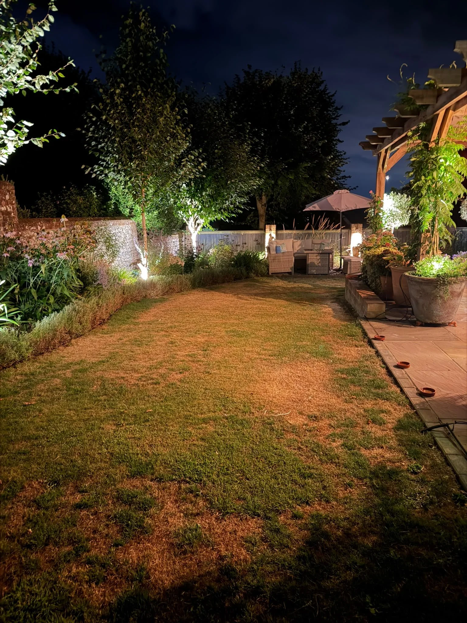 Nighttime backyard with lit trees, garden flowers, patio furniture, and an umbrella, illuminated by outdoor lighting.