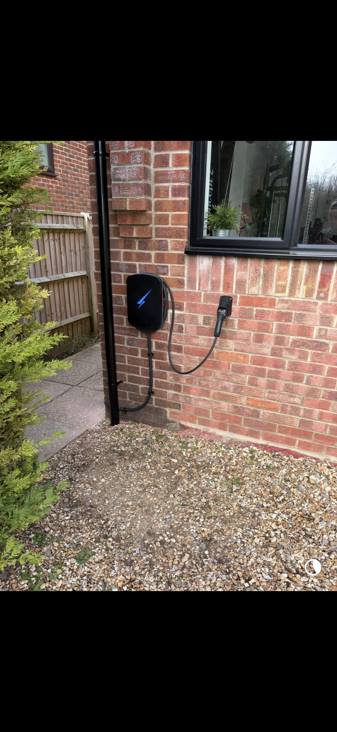 Electric vehicle charging station installed on a brick wall outside a house, with charging cable and connector, near a window and garden.