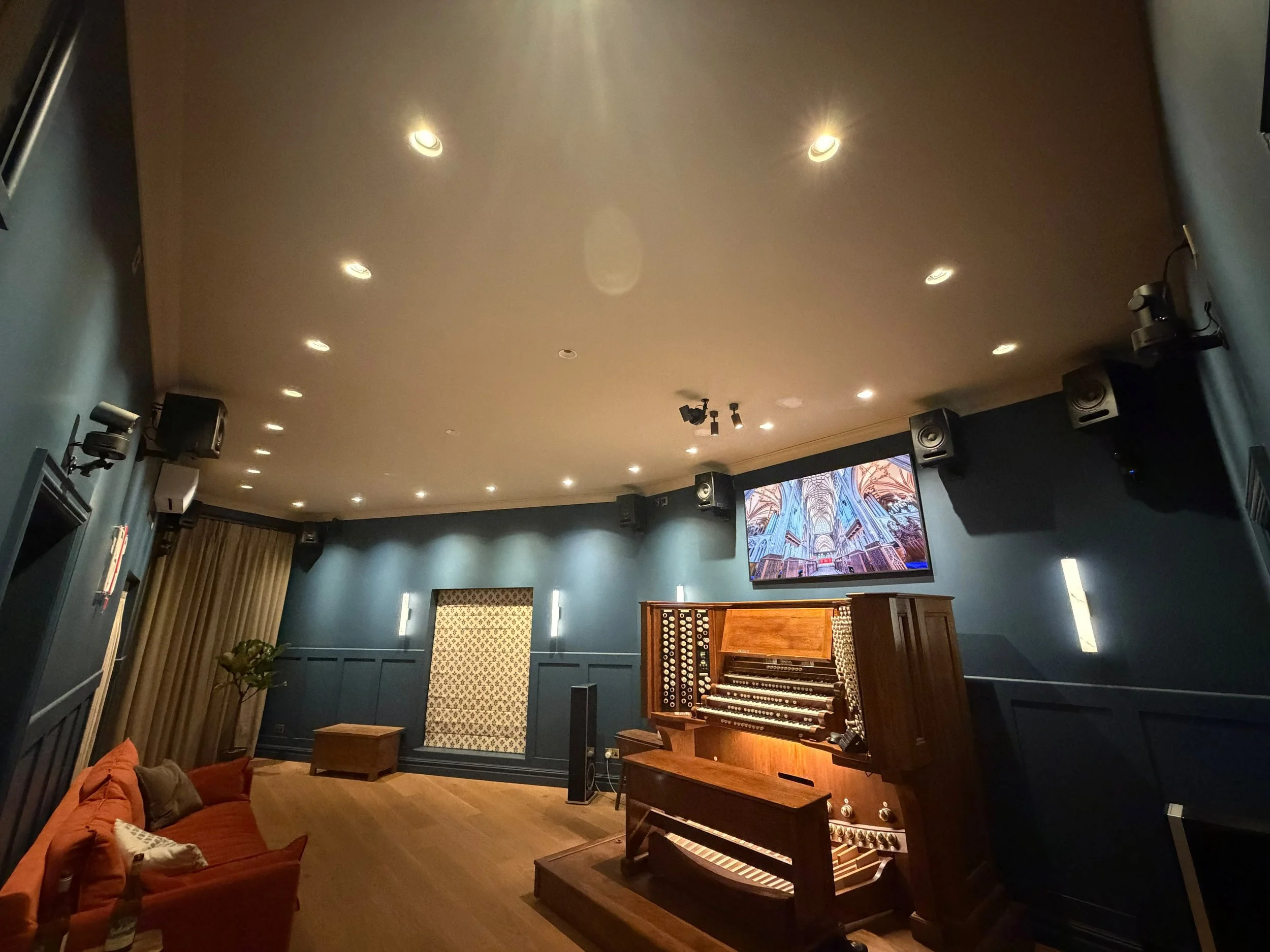 A music room with a wooden pipe organ on a wooden stage, an orange velvet sofa with pillows, a large screen, and multiple speakers and ceiling lights.