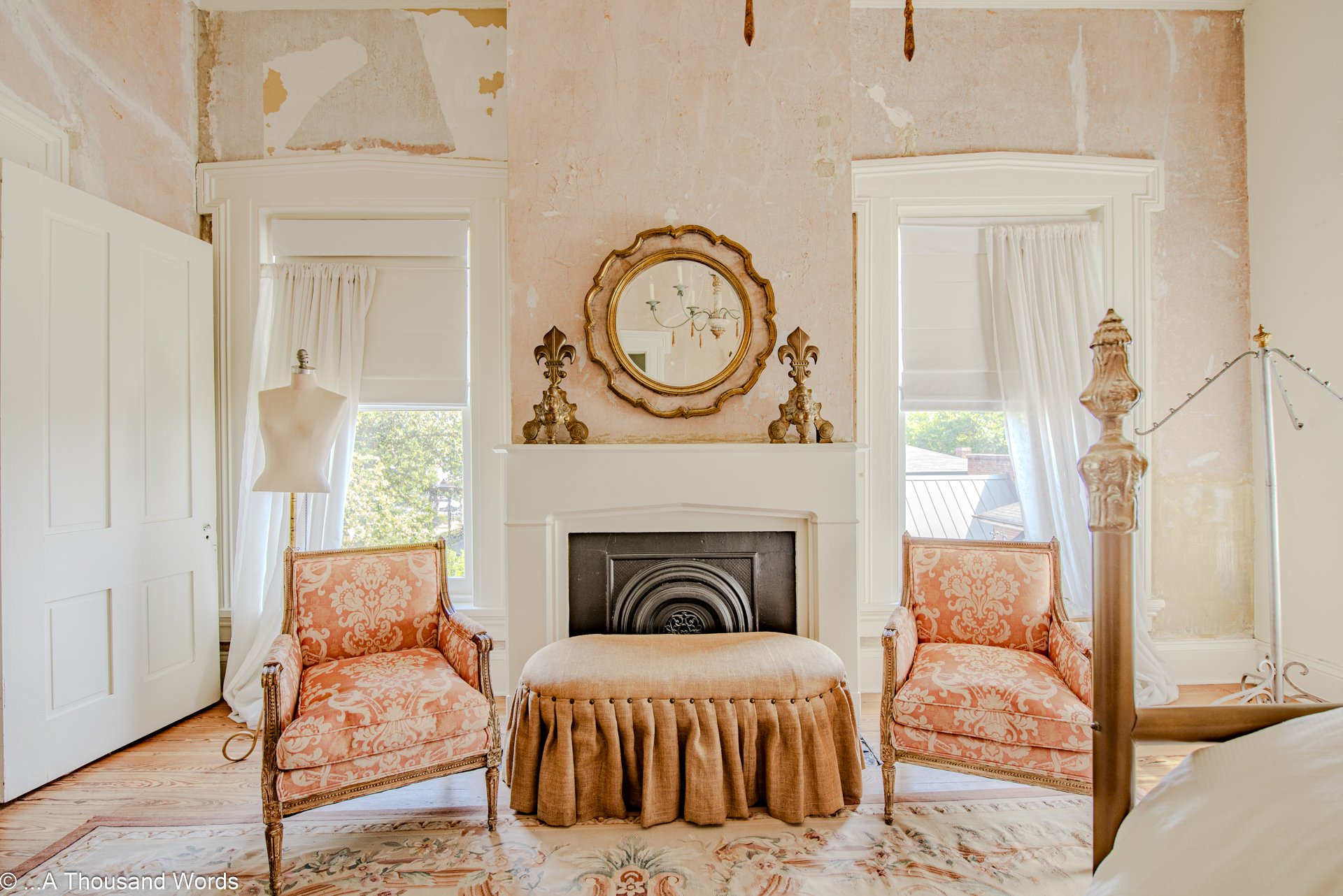 A vintage living room with two pink patterned armchairs, a pink ottoman, a white fireplace with a mirror above, brass candlesticks, two windows with white curtains, and a chandelier reflected in the mirror. Auburn real estate photoshoot. Airbnb, VRBO