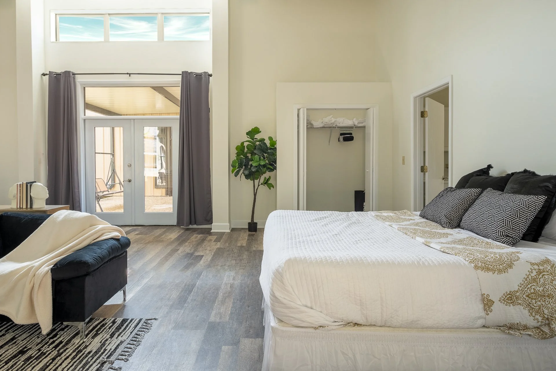 A bedroom with a white bed, black pillows, and a decorative bedspread. There is a black bench with a blanket and a striped rug. Real estate listing in Auburn, AL