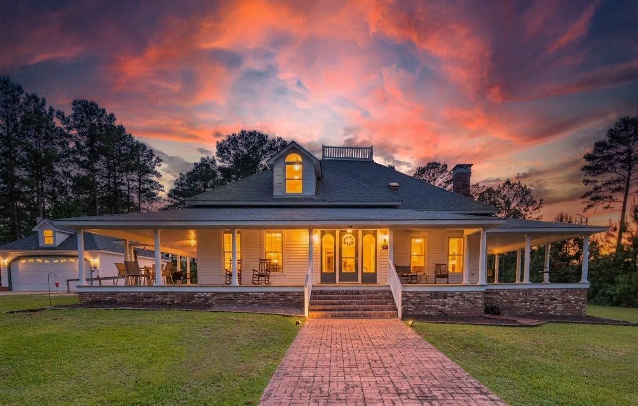 A large house with a wrap-around porch illuminated from inside, set against a sunset sky with pink and orange clouds. The house has a brick walkway leading to steps, a front door with glass panels, and trees in the background. Twilight photography.