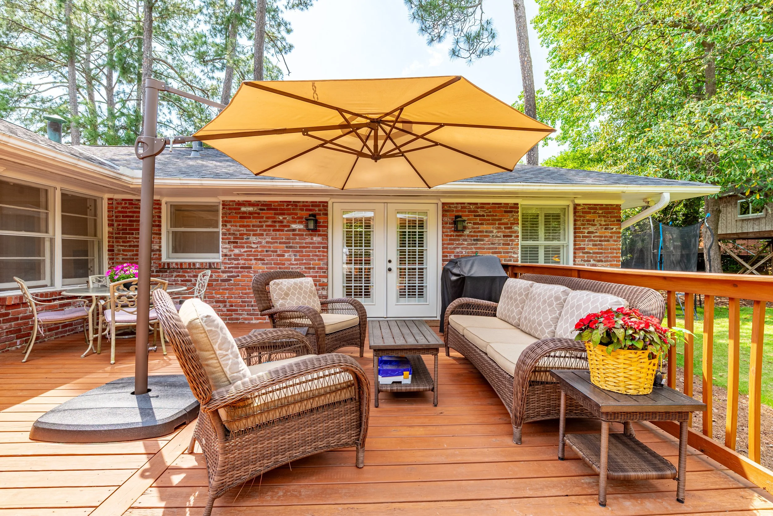 A wooden backyard deck with outdoor furniture, including wicker chairs and a bench with cushions, and a large yellow patio umbrella. There are potted flowers, a grill, and a glass door leading into a brick house. Hilton Heights neigborhood.