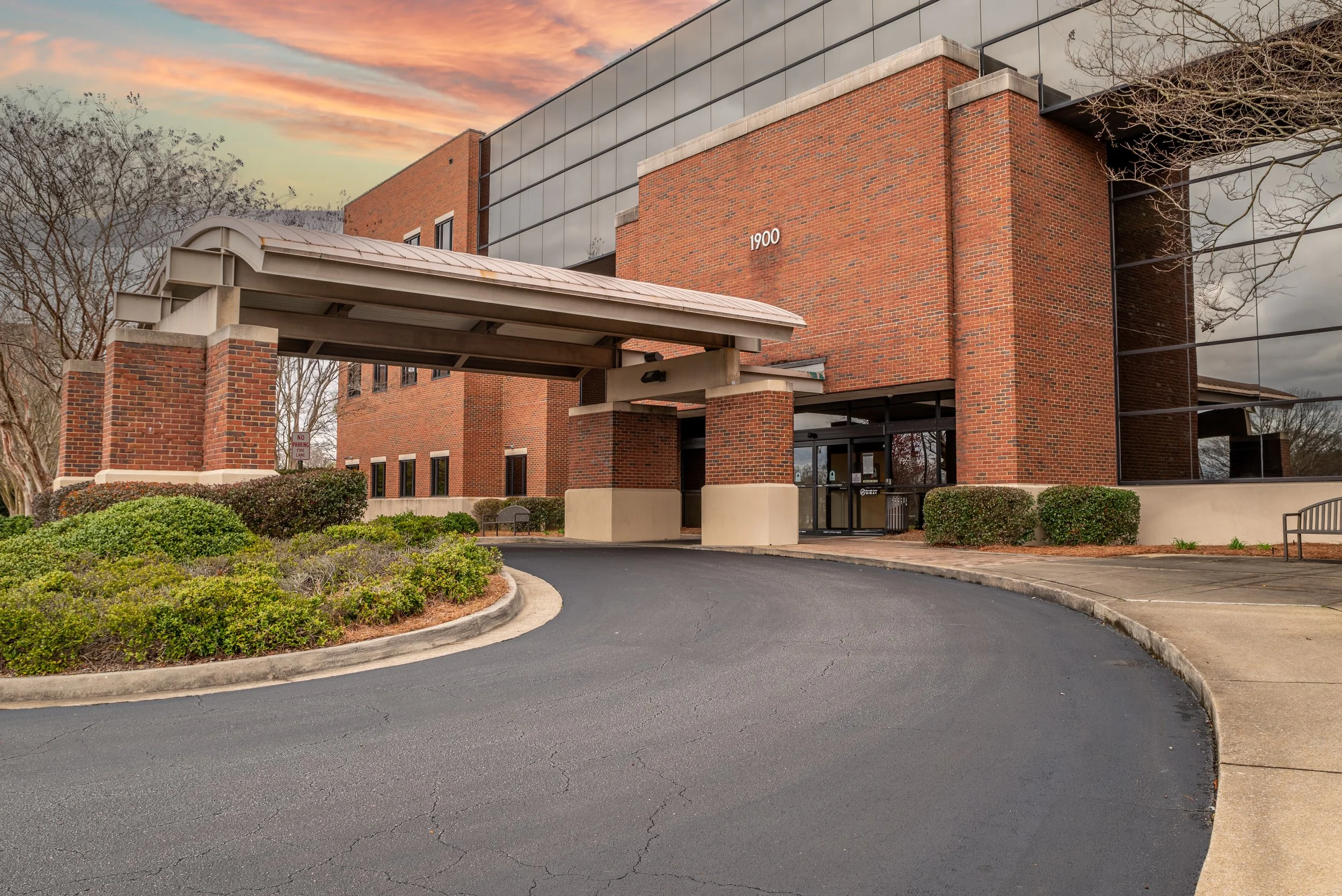 Exterior of a modern brick building with a curved metal awning at the entrance, surrounded by landscaped bushes and trees with a colorful sunset sky in the background.