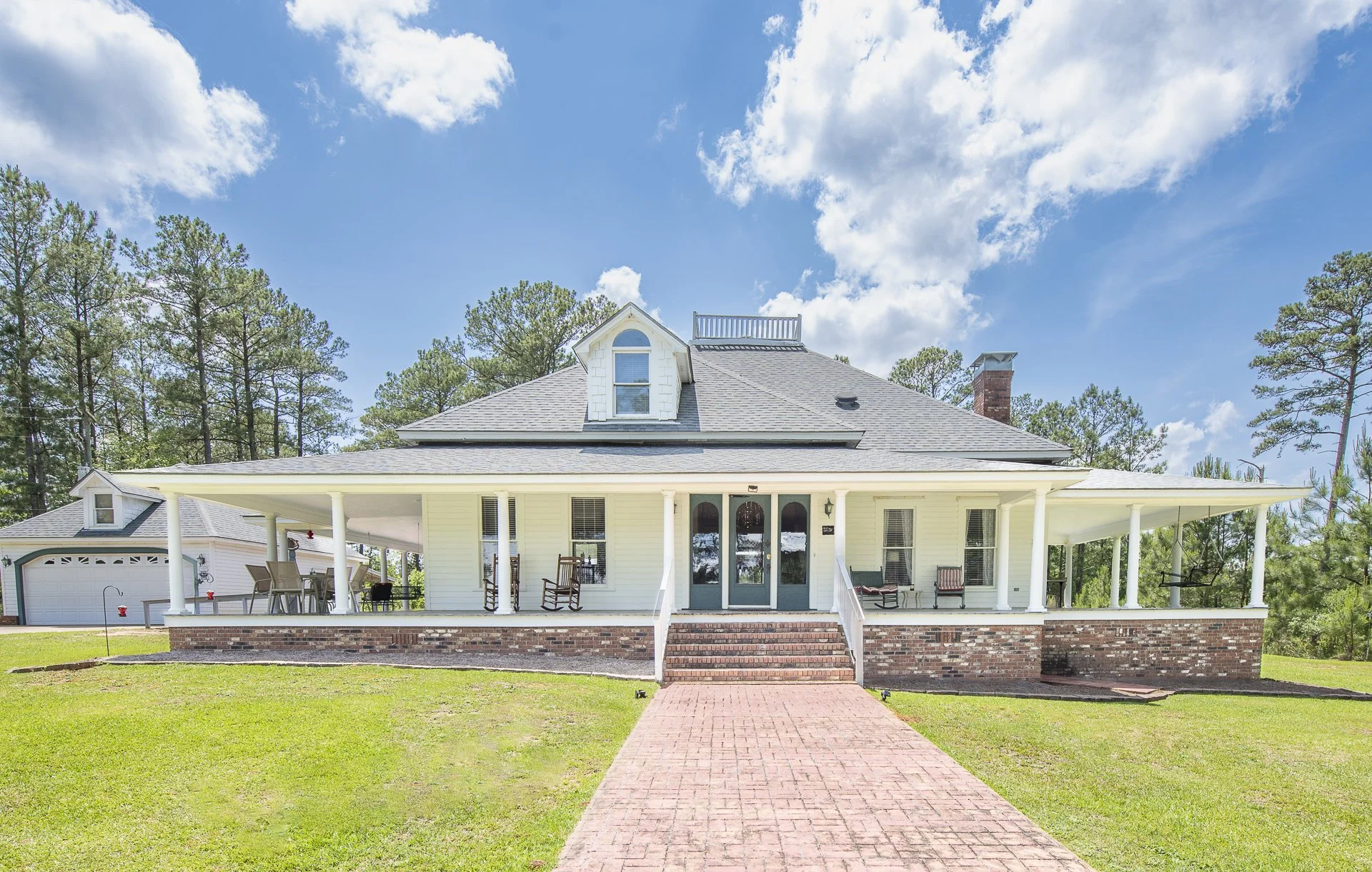 Front view of a two-story white house with a large wrap-around porch, brick steps leading to the entrance, surrounded by green lawn, tall trees, blue sky, and some clouds. New listing in Opelika, AL area.
