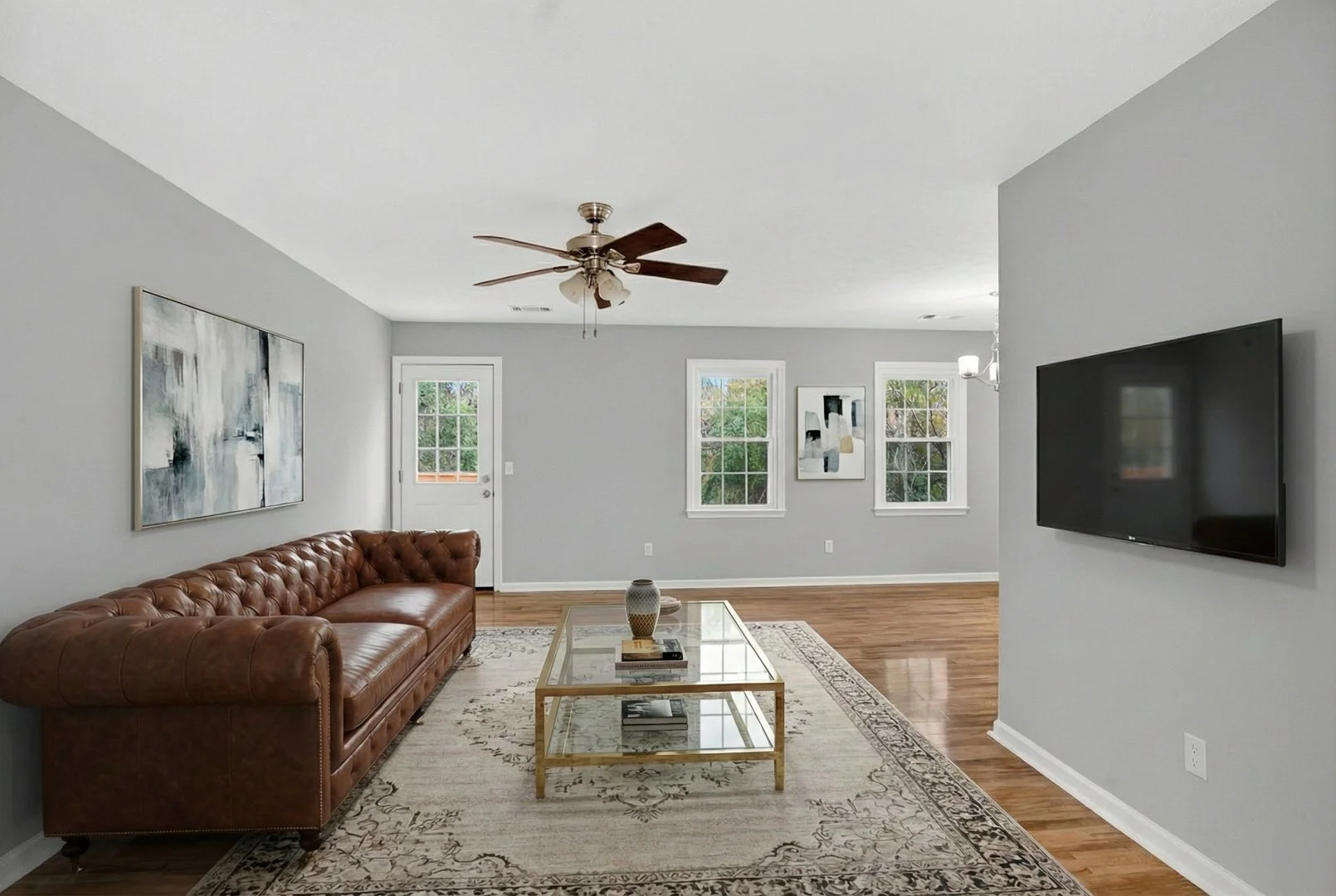 Living room with a leather couch, glass coffee table, wall-mounted TV, paintings, windows, and hardwood floor. Virtual Staging. Columbus, GA. Real estate listing