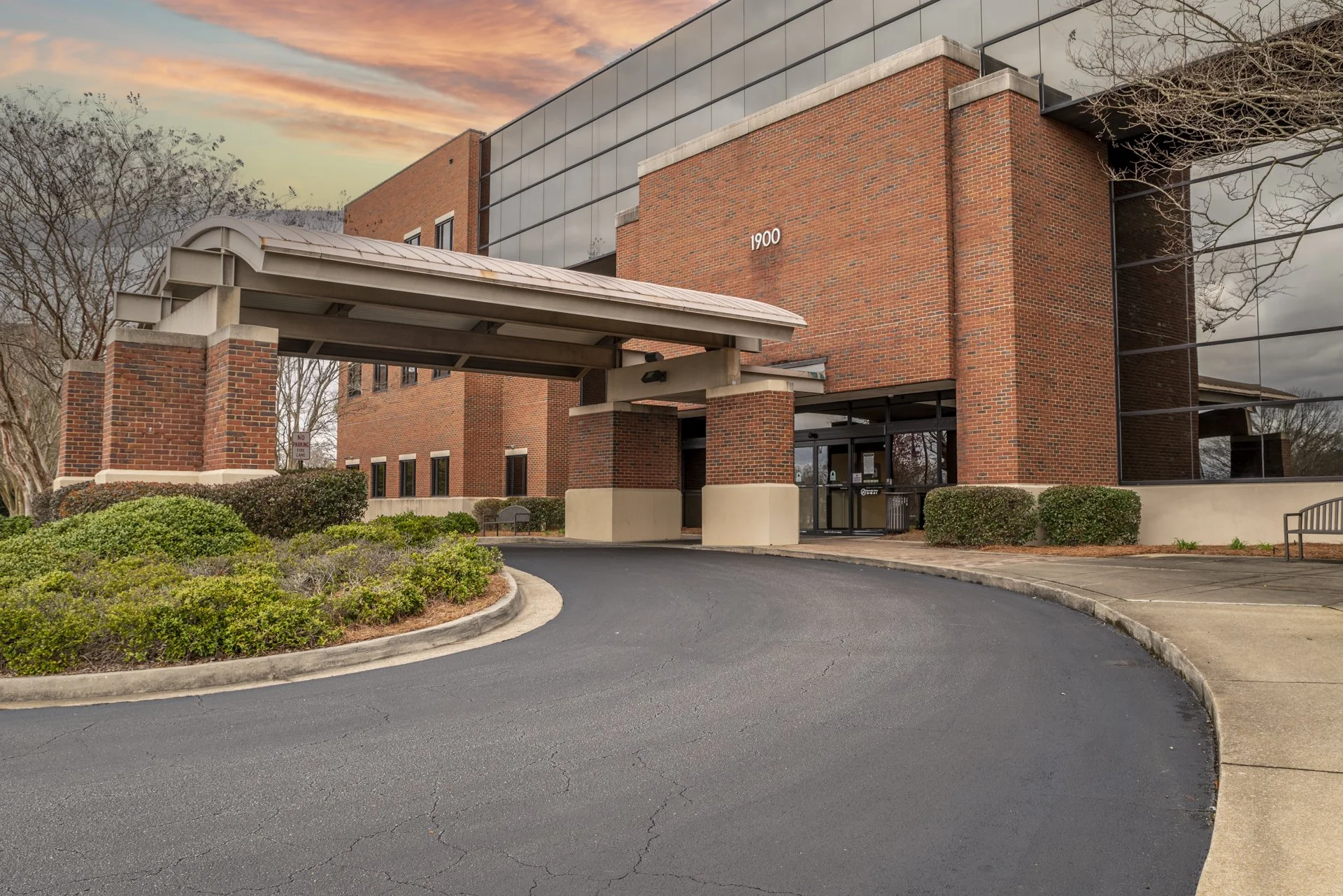 A modern brick office building with reflective glass windows at sunset, surrounded by landscaped bushes and a paved parking lane. Talbotton Road medical office photoshoot. Architectural photography.