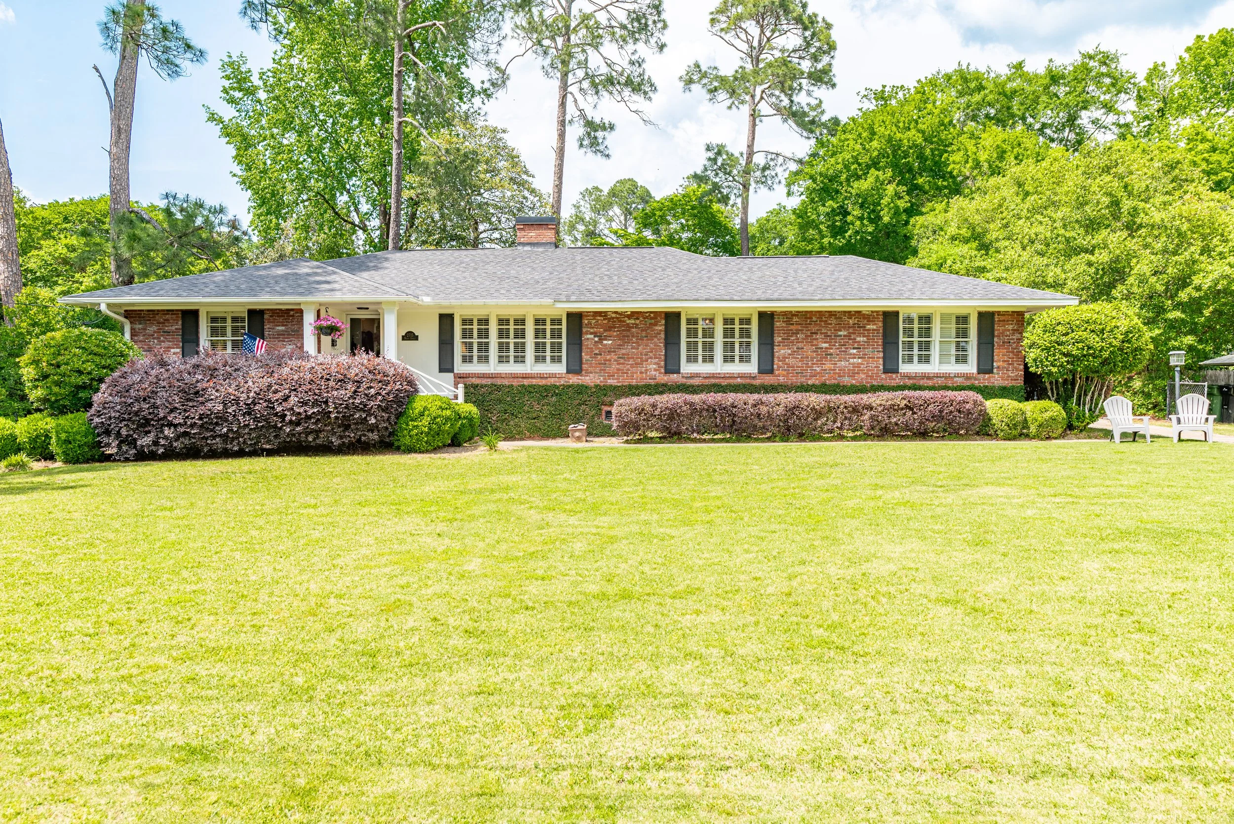 A single-story brick house with white trim, surrounded by a lush green lawn and tall trees, featuring a front porch with hanging flower baskets and a small American flag near the entrance.