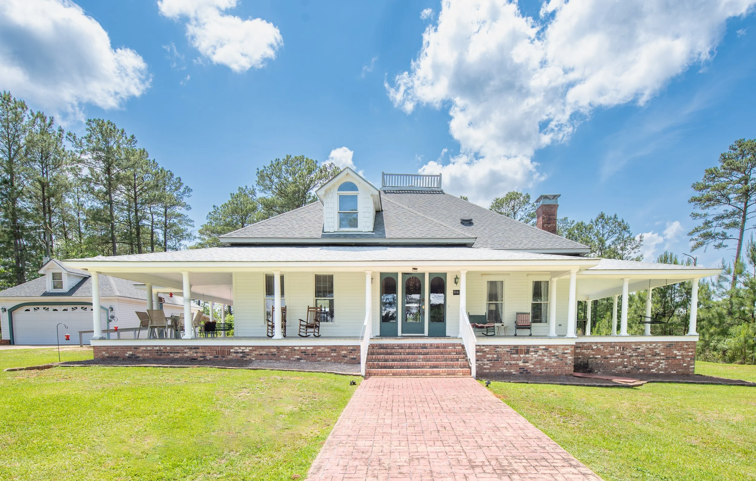 White house with a large porch, multiple rocking chairs, brick steps, and a brick walkway leading to the house, with trees and a bright cloudy sky in the background.