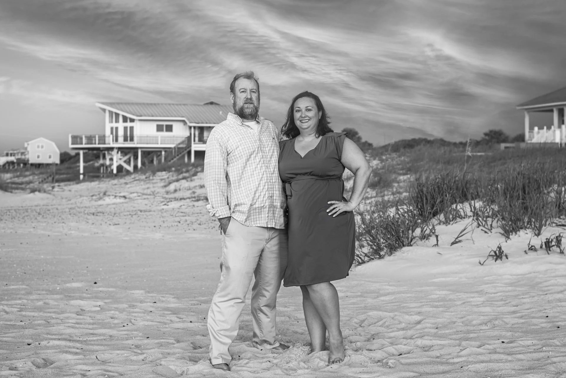 A black and white photo of a man and woman standing on a sandy beach. The man has a beard, is wearing a checked shirt and light-colored pants. The woman is wearing a dark dress and has her hand on her hip, smiling. In the background, there are beach houses on stilts and a sky with wispy clouds.