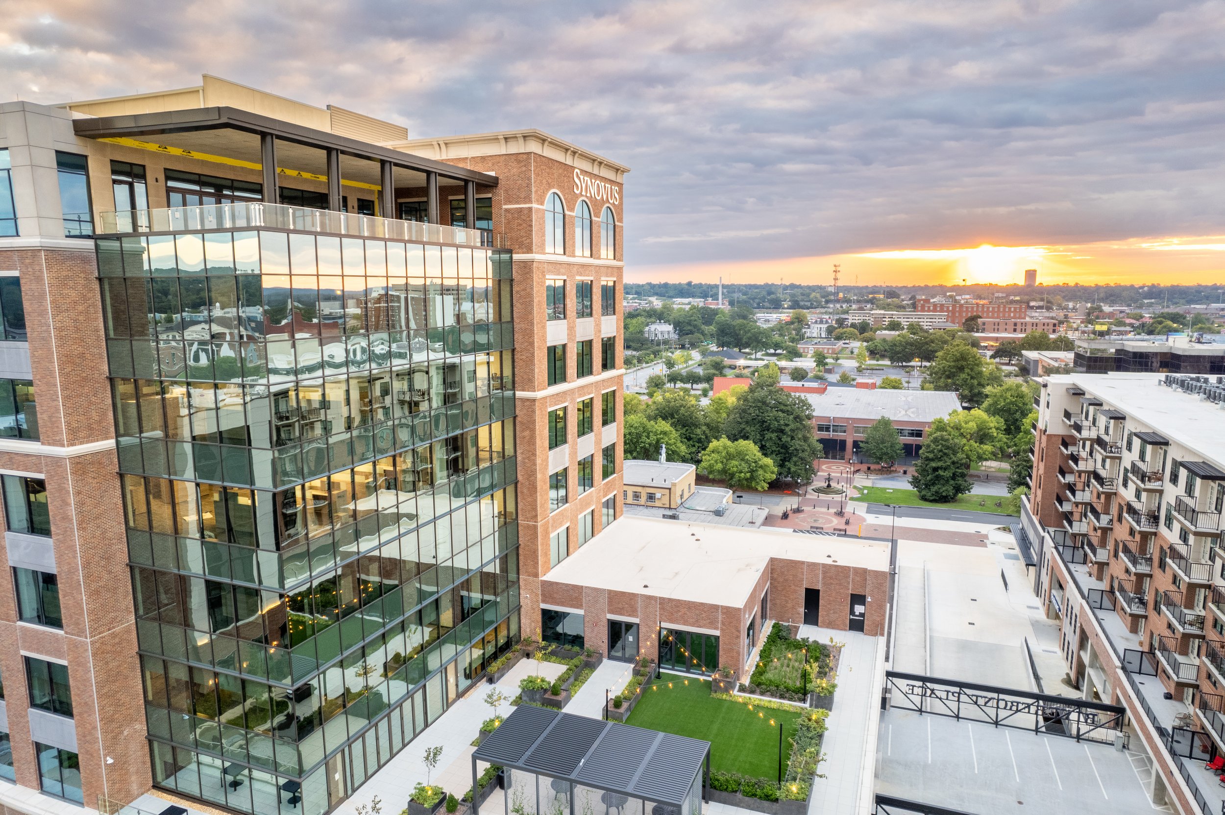 A modern multi-story building with a glass facade reflecting other buildings and the sky, a small outdoor garden with string lights, and a cityscape with trees and additional buildings under a partly cloudy sky at sunset.