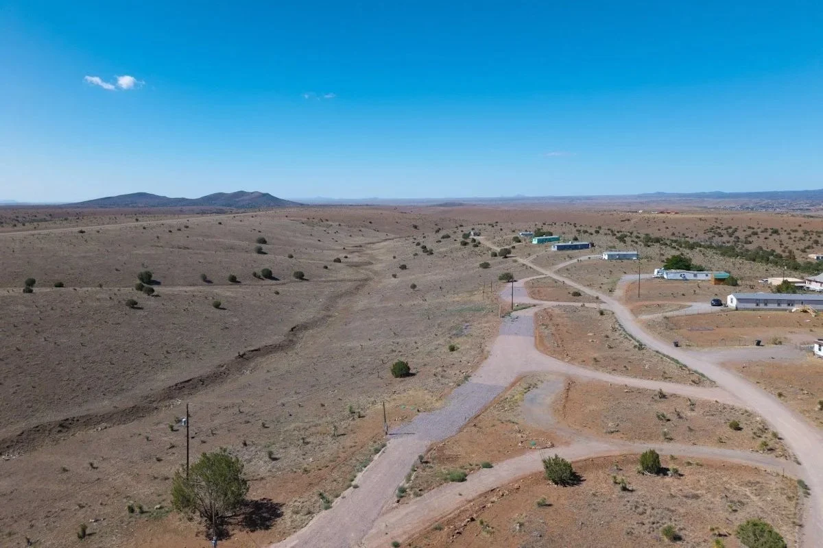 A desert landscape with scattered small bushes and a few buildings in the distance, under a clear blue sky.