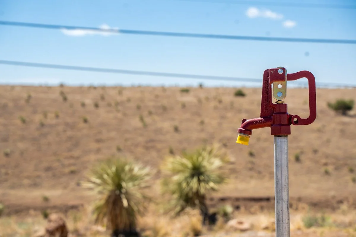 Red garden hose faucet in desert landscape with sparse vegetation and power lines in the background.