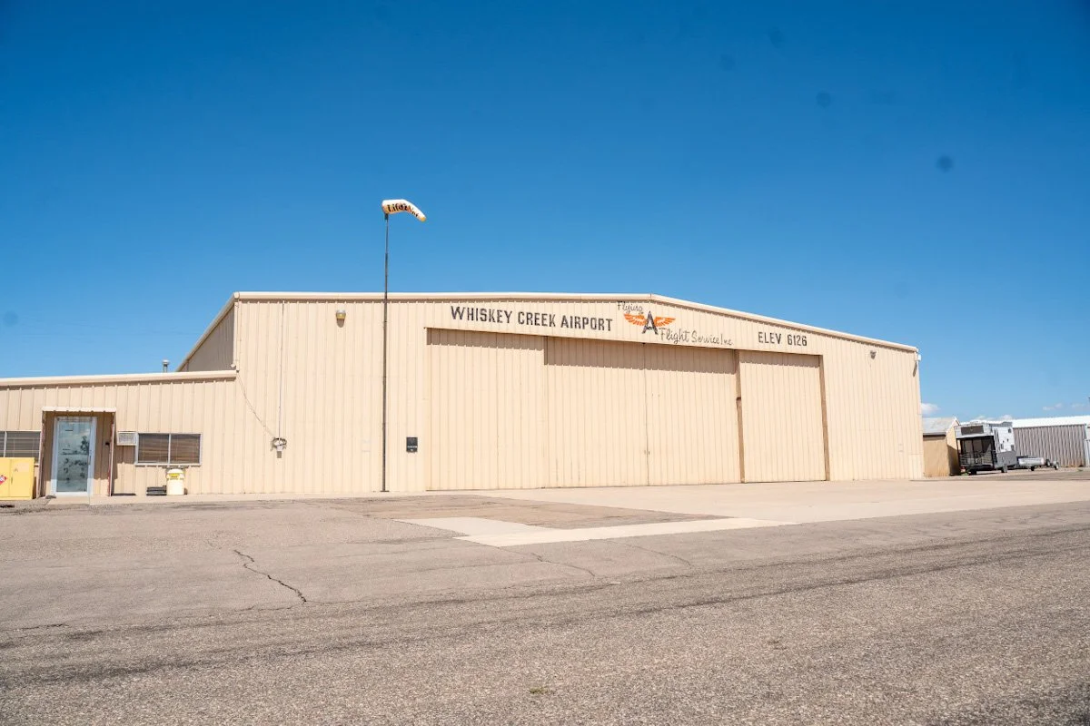 Exterior view of Whiskey Creek Airport building with a beige metal facade, a small door and windows to the left, a flag on a pole, and a trailer in the background under a clear blue sky.