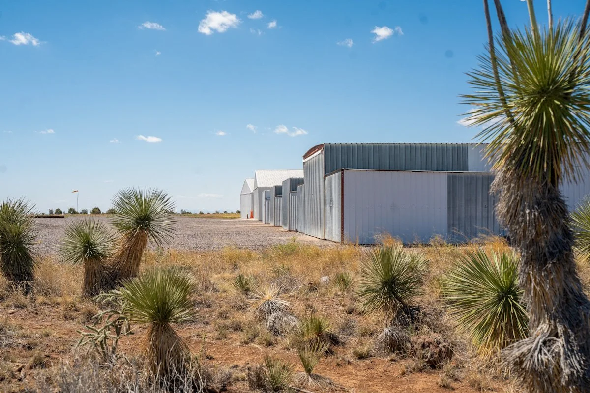 A row of metal buildings in a desert landscape with dry grass and Joshua trees under a blue sky.