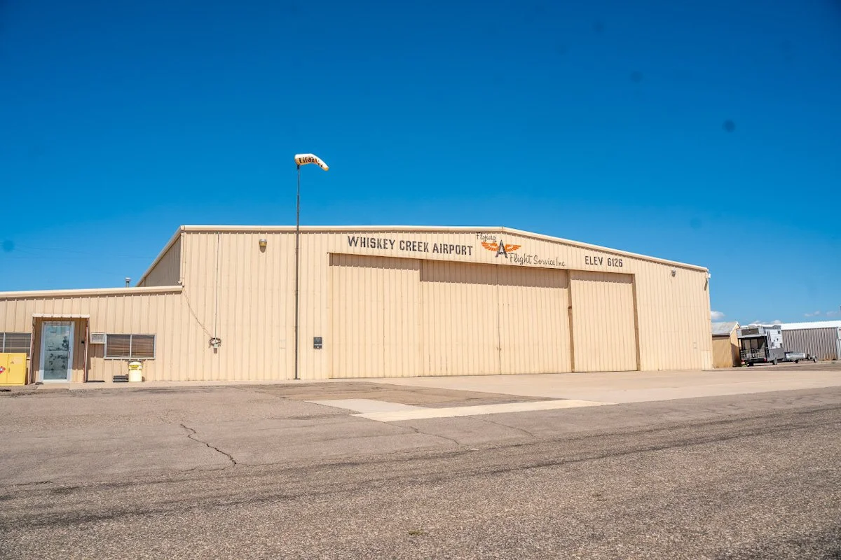 Small beige building with a sign that reads Whiskey Creek Airport, located at elevation 6126 feet, under a clear blue sky.