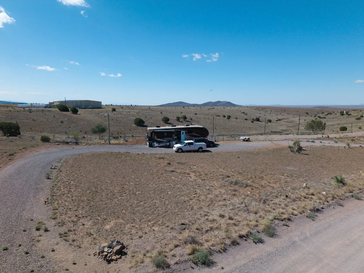 An RV parked at Whiskey Creek Mobile Estates on a gravel lot in a New Mexico landscape with sparse vegetation, small bushes, and a few distant hills under a bright blue sky.