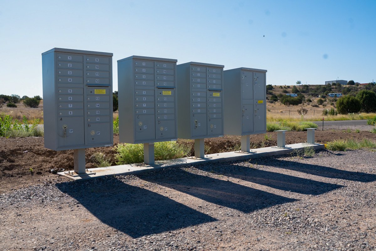 A row of gray mailbox lockers standing on a concrete pad in an outdoor rural area with sparse vegetation and a clear blue sky.
