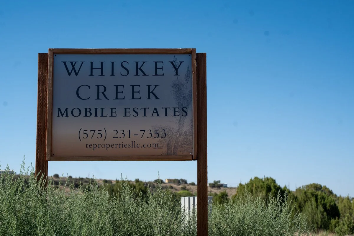 Sign for Whiskey Creek Mobile Estates with contact number and website, surrounded by shrubs and clear blue sky.