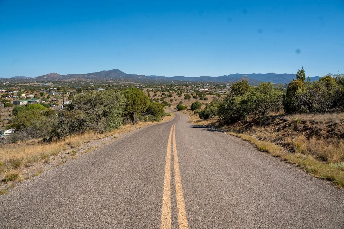 A two-lane road curves through a dry, hilly landscape with scattered trees and shrubs under a clear blue sky, with mountains in the distance.
