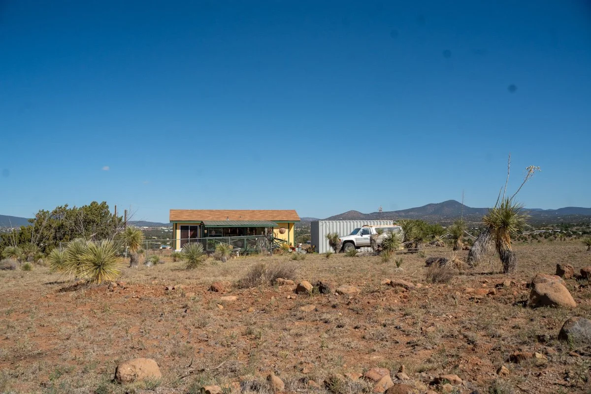A small house with a porch, a pickup truck, and a large white storage container in a desert landscape with sparse vegetation and mountains in the background under a clear blue sky.