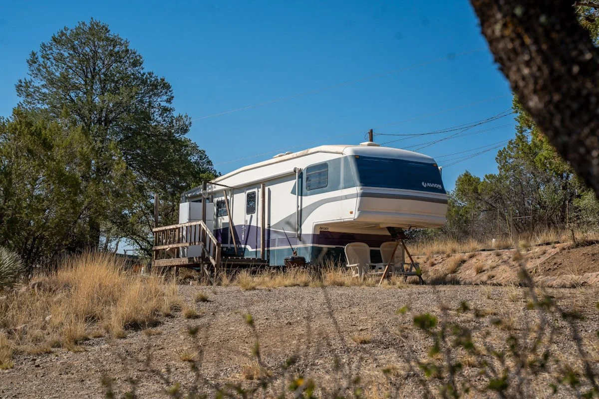 A travel trailer parked in a dry, grassy outdoor area with trees and a blue sky in the background.