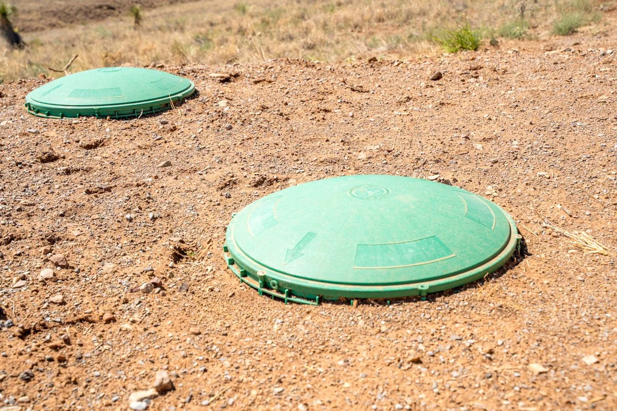 Two green underground utility access covers on sandy ground in an open outdoor area.