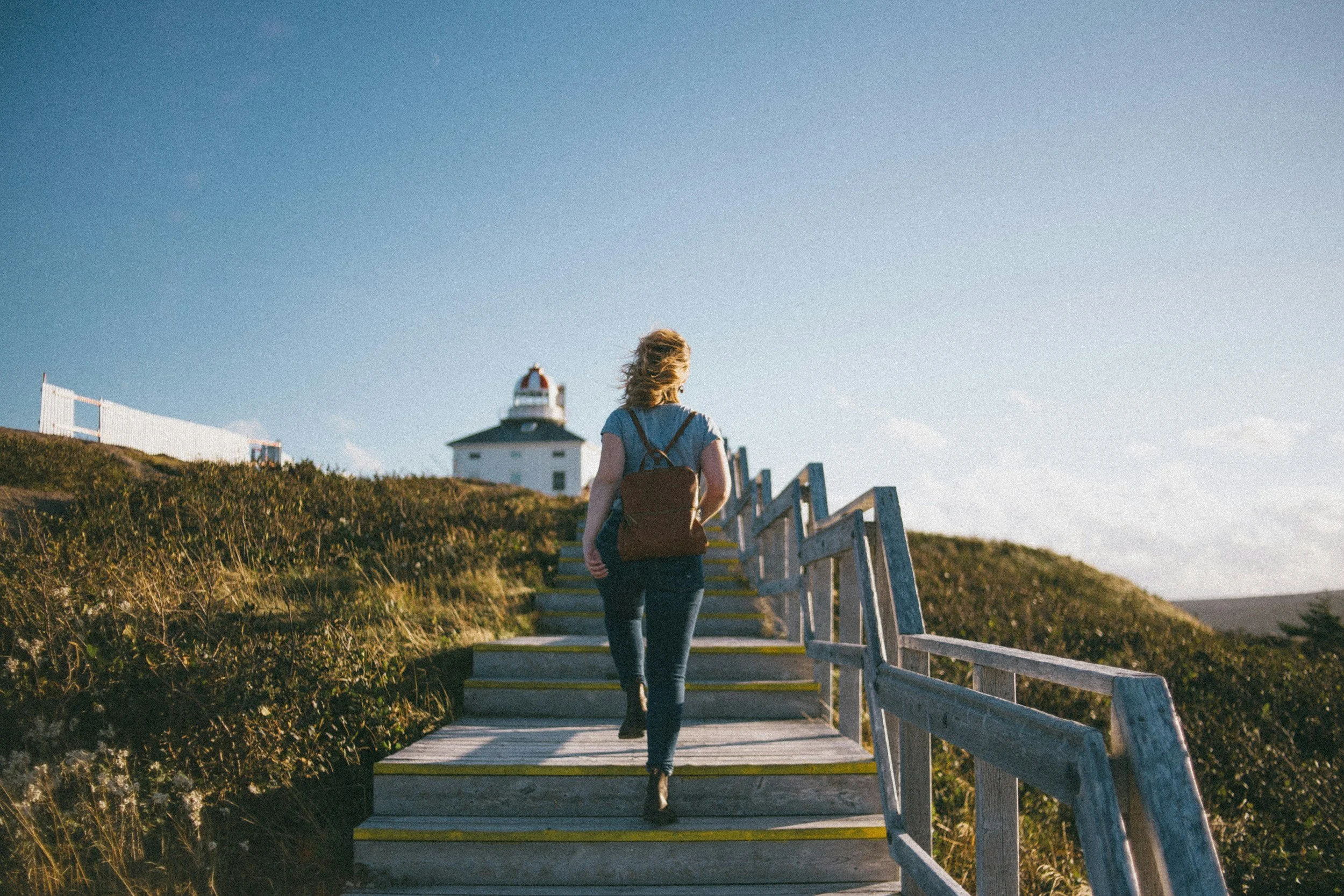 A woman with blonde hair walking up wooden stairs towards a lighthouse under a clear blue sky.