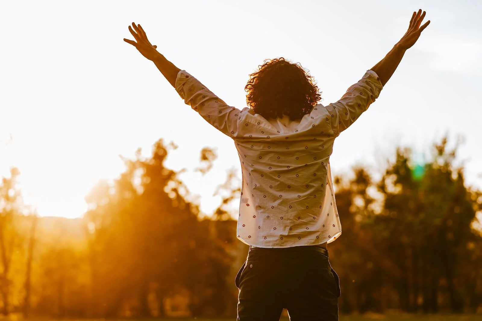 Person with curly hair raising arms in the air during sunset outdoors, with trees in the background.