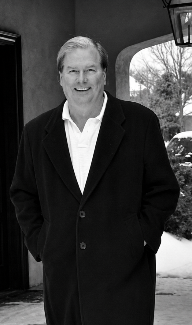 A smiling middle-aged man with light hair, wearing a dark overcoat and a white shirt, standing outdoors near a building with a window and archway, with trees and parked cars in the background.