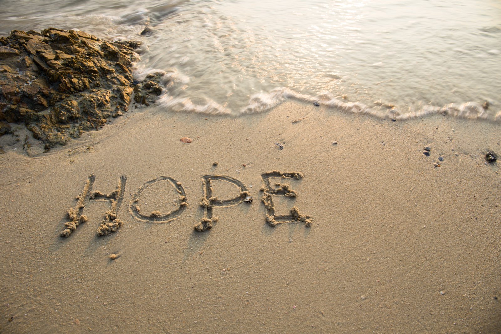 The word 'HOPE' written in the sand on a beach, with the ocean and waves lightly touching the shore in the background.