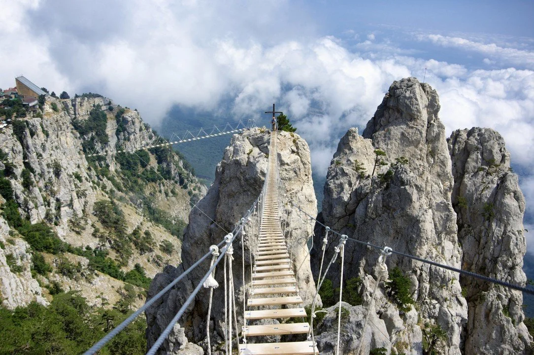 A narrow suspension bridge with wooden planks and metal ropes crossing between large rocky peaks in a mountainous area under cloudy sky.