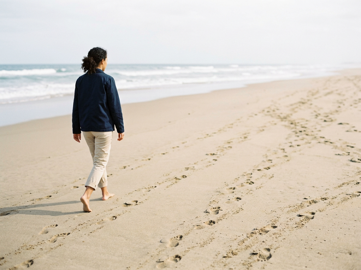 A woman with curly hair walking barefoot along a sandy beach with footprints, facing the ocean under an overcast sky.