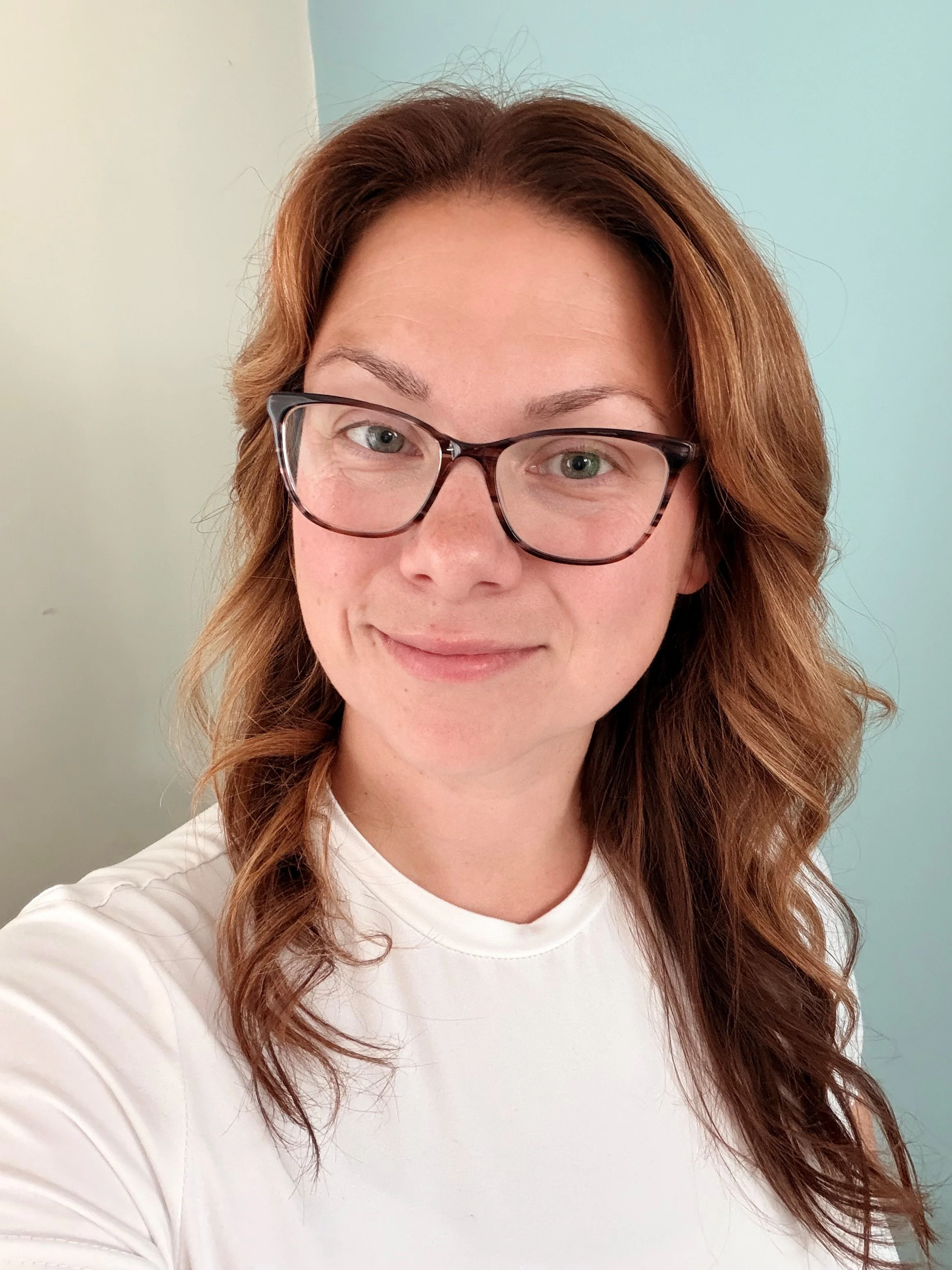 A woman with light skin and red hair styled in loose waves, wearing glasses and a white shirt, taking a selfie in front of a light-colored wall.