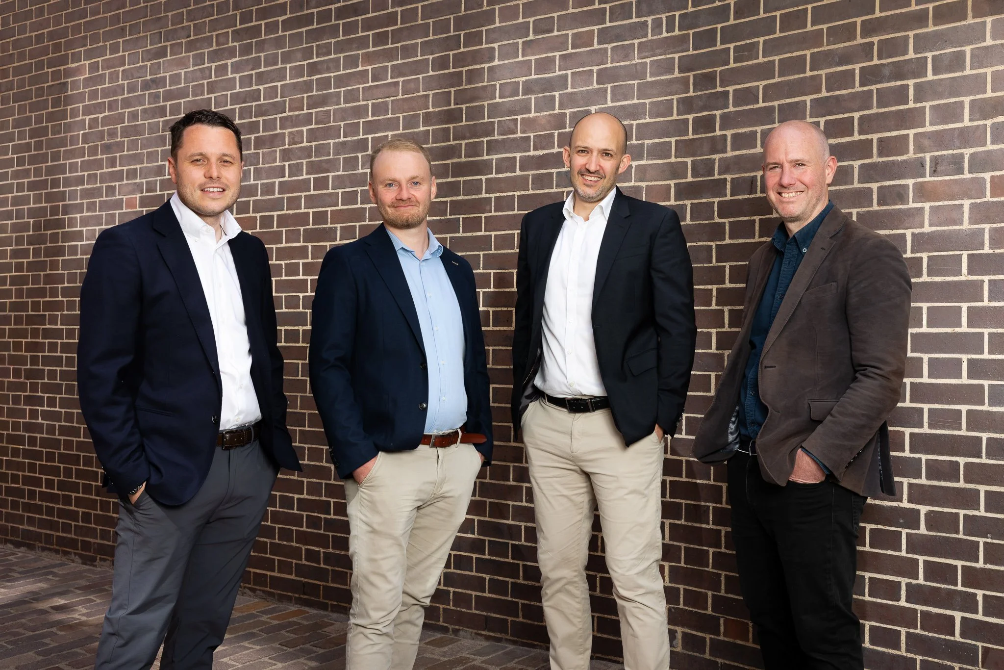 A group of four men standing in front of a brick wall, dressed in business casual and formal attire, smiling at the camera.