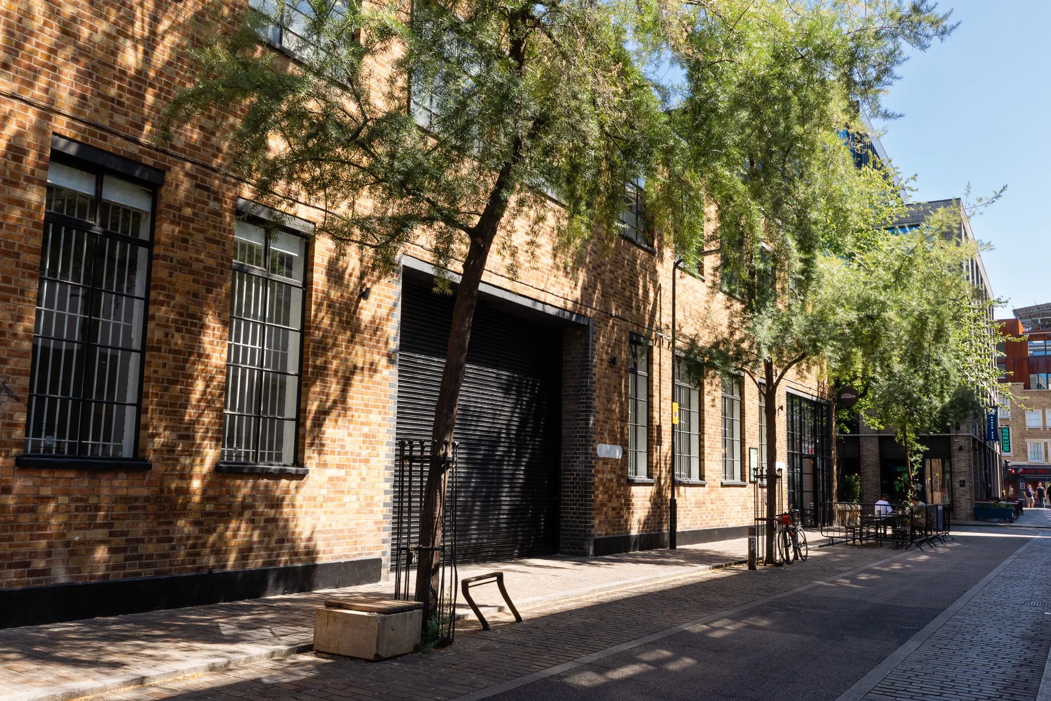 A city street scene with brick buildings, trees casting shadows, and a sidewalk with benches and bicycles. People are visible in the background.