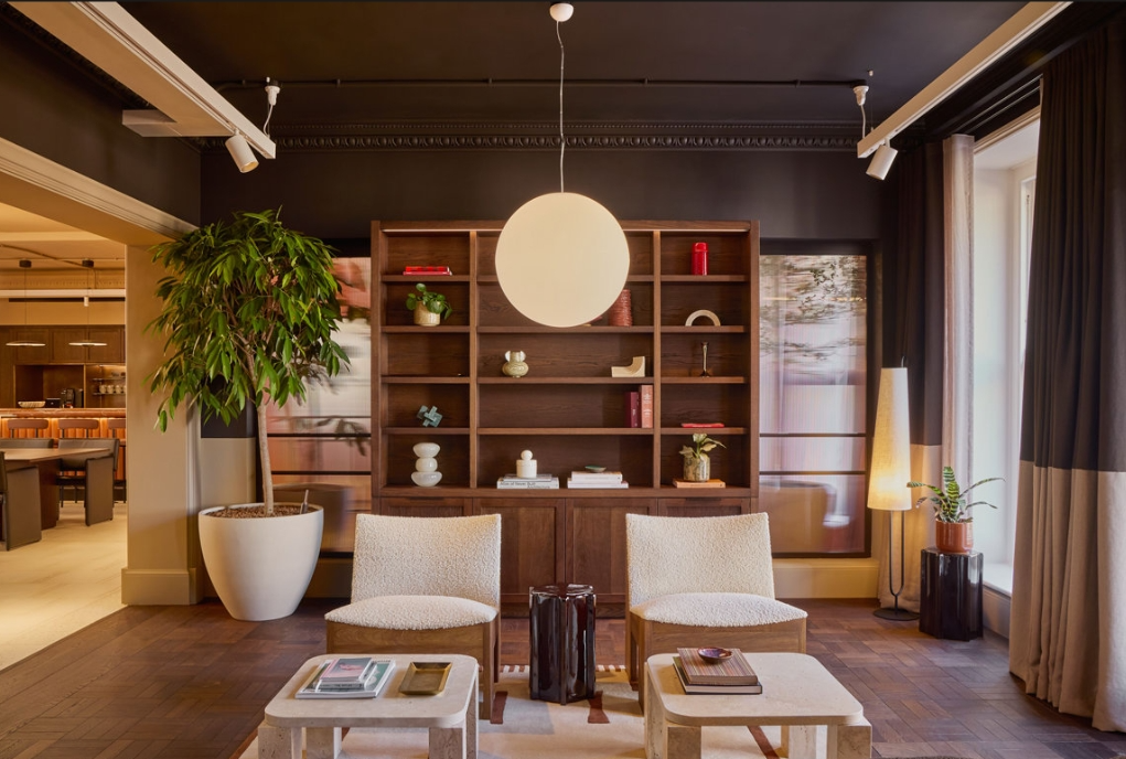 A cozy living room with two beige chairs, a white coffee table with magazines, a tall potted plant, a wooden bookshelf with decorative items, and a floor lamp near a window with curtains.