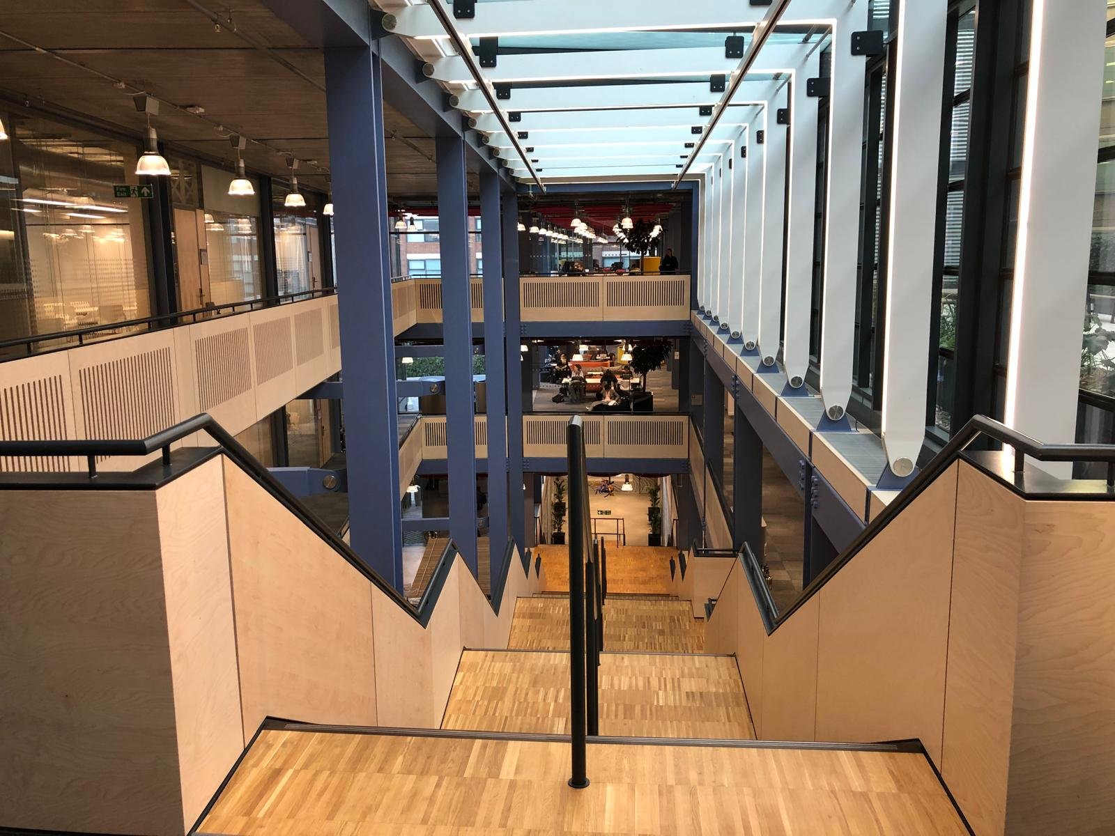 Multi-level modern interior with staircase and walkways, glass ceiling, and people working or socializing in the background.