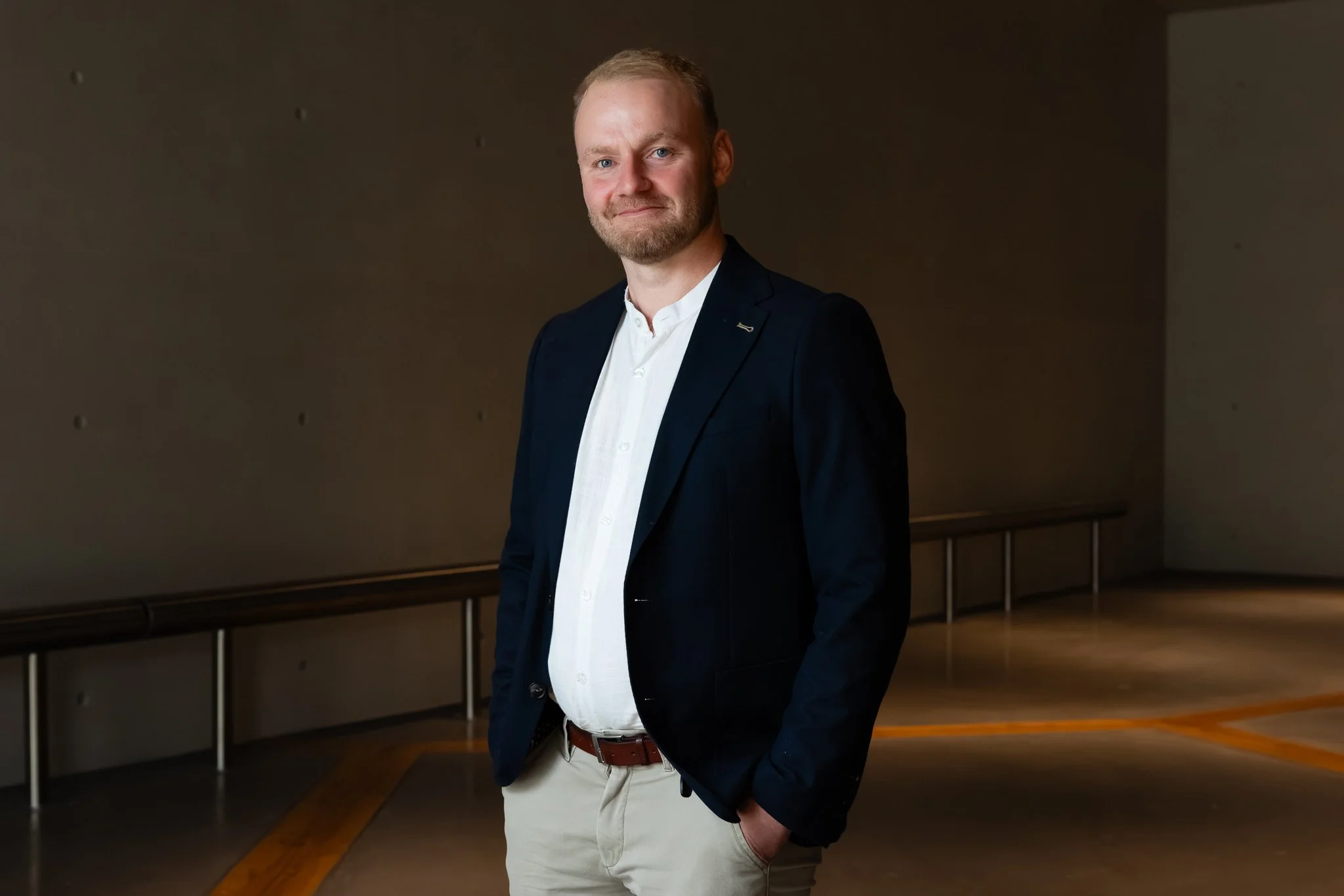 A man in a dark blazer, white shirt, and khaki pants standing in an indoor area against a plain wall.