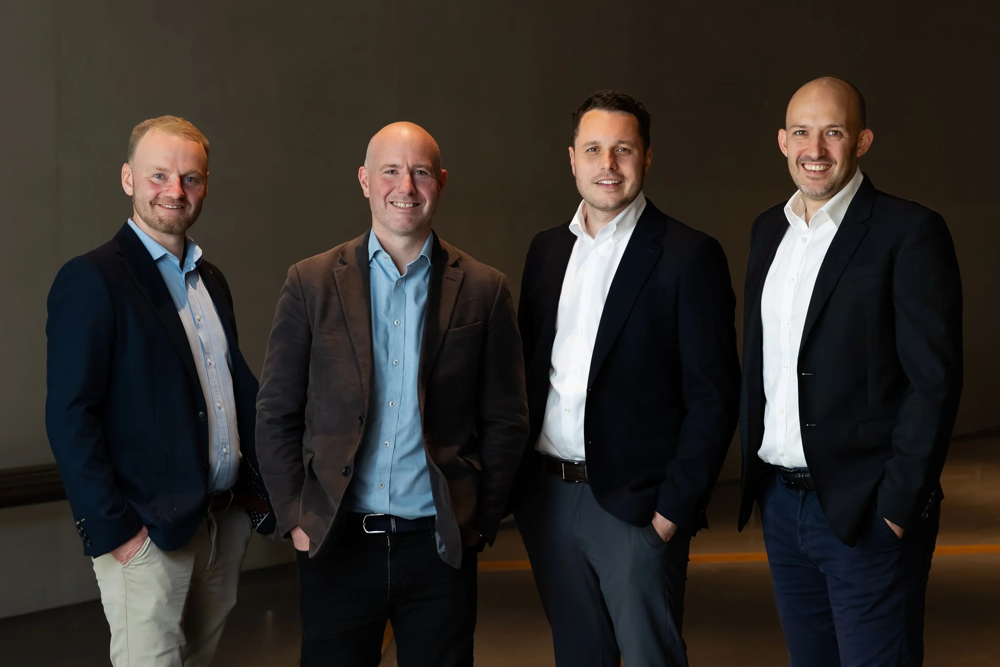 Four men in business attire standing together indoors against a dark background, smiling at the camera.
