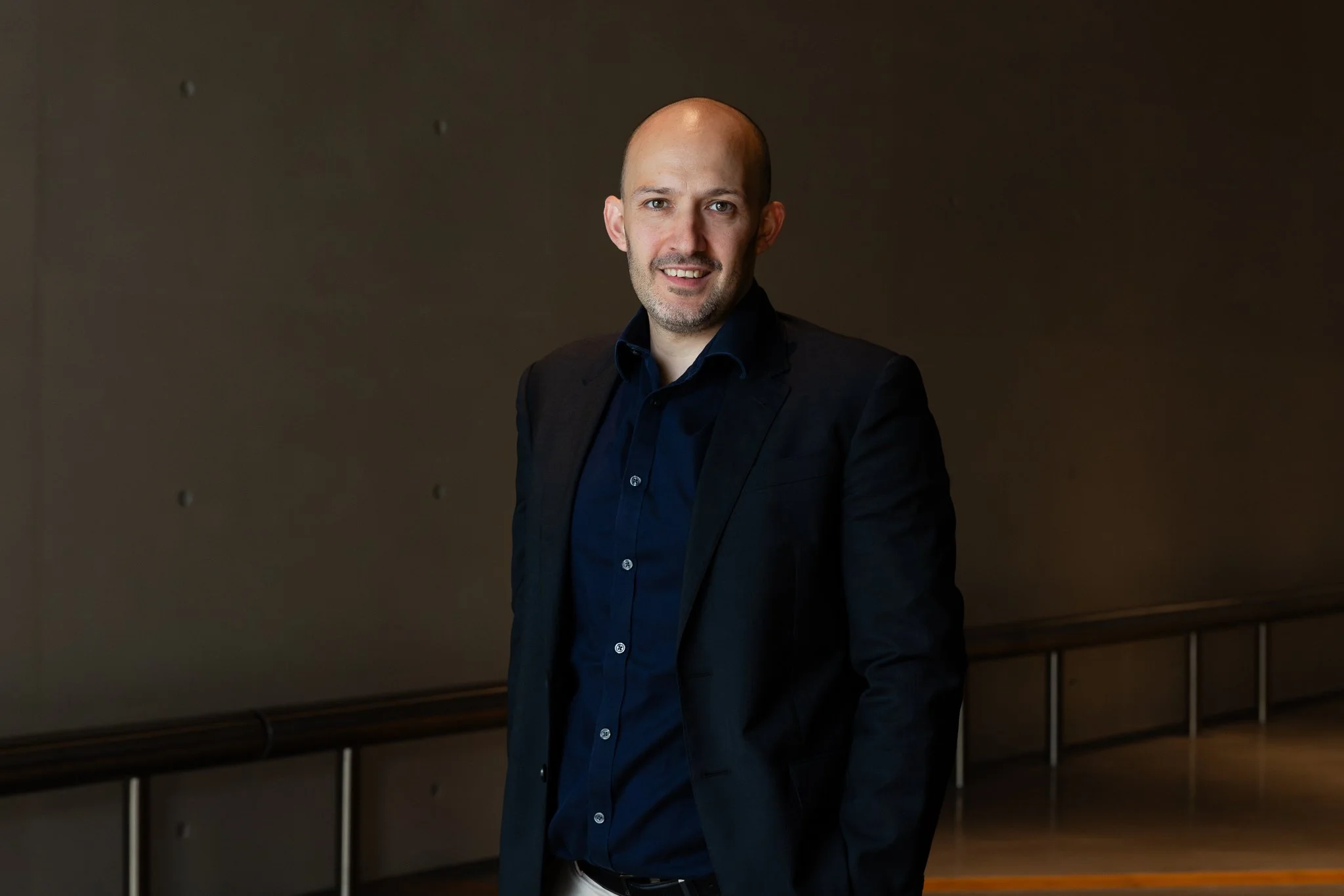 Bald man in black suit and navy shirt standing indoors against a dark background, smiling slightly, with his hands in his pockets.