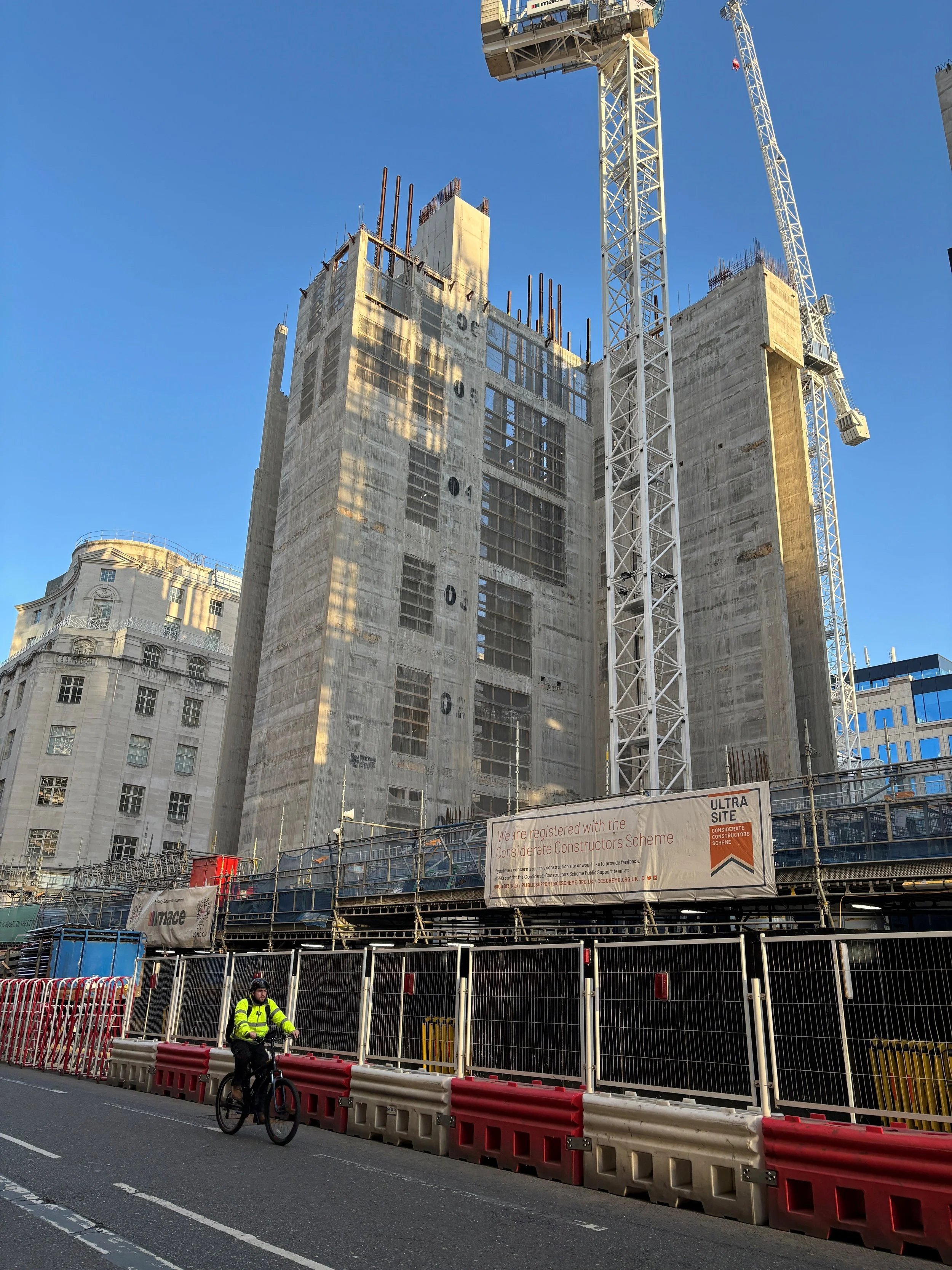 A construction site with a tall building under construction, cranes, and a construction worker riding a bicycle in the foreground. Safety barriers and signage are also visible.
