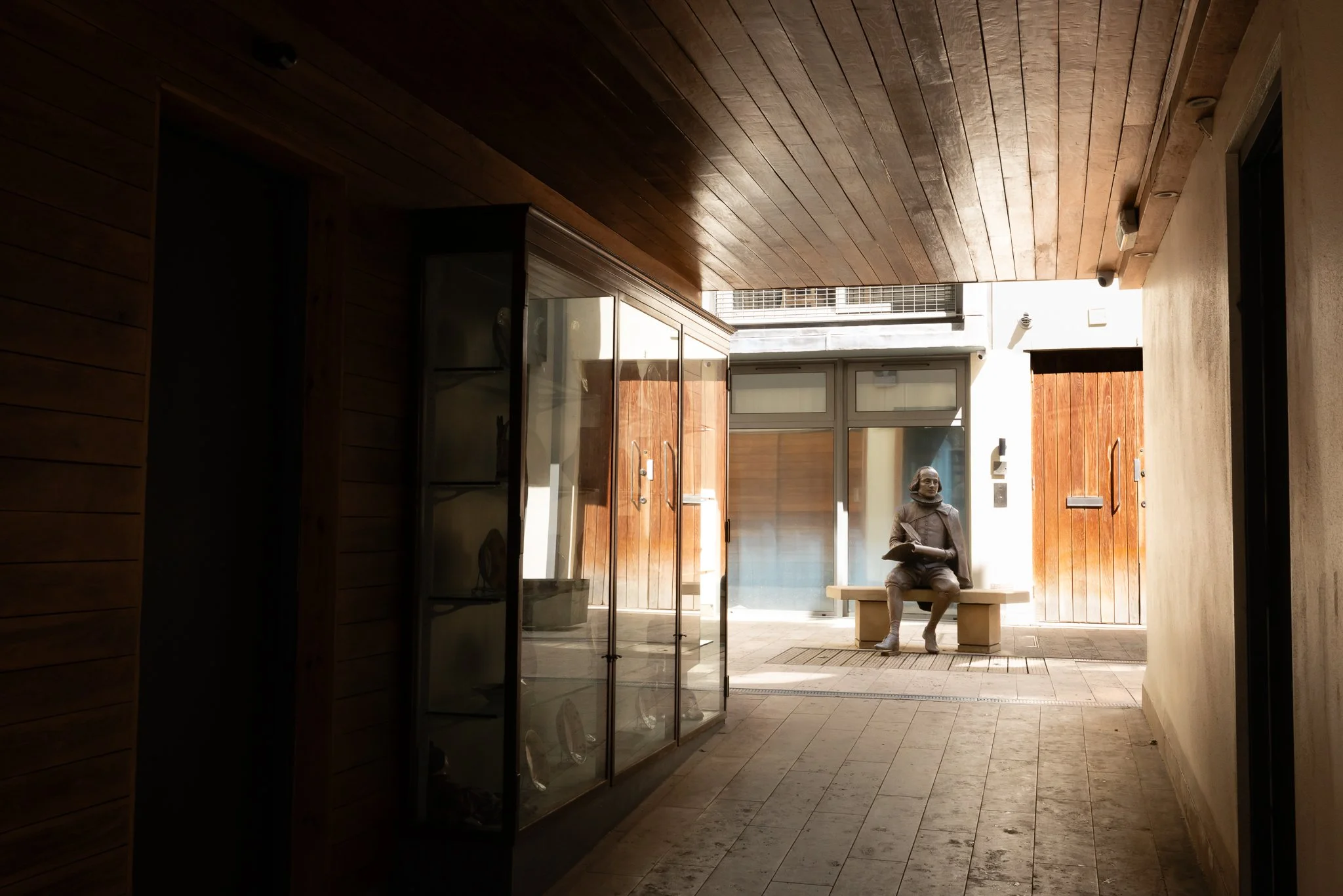 Interior view of a building with a wooden ceiling, tiled floor, and glass doors leading to an outdoor area with a sculpture of a person seated on a bench holding a book.