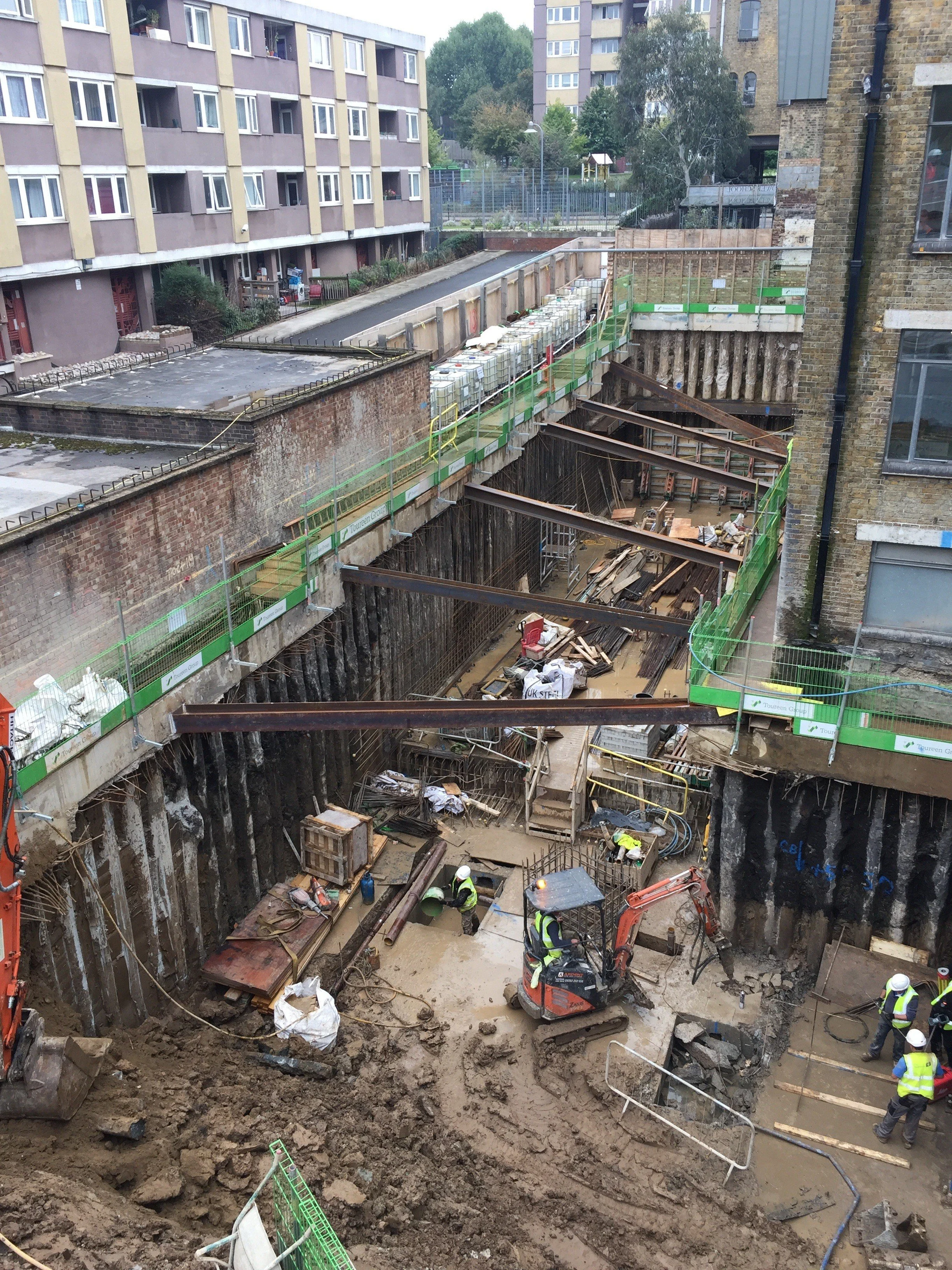 Construction workers wearing safety helmets and reflective vests working in an underground excavation site surrounded by scaffolding and building structures, with residential buildings in the background.