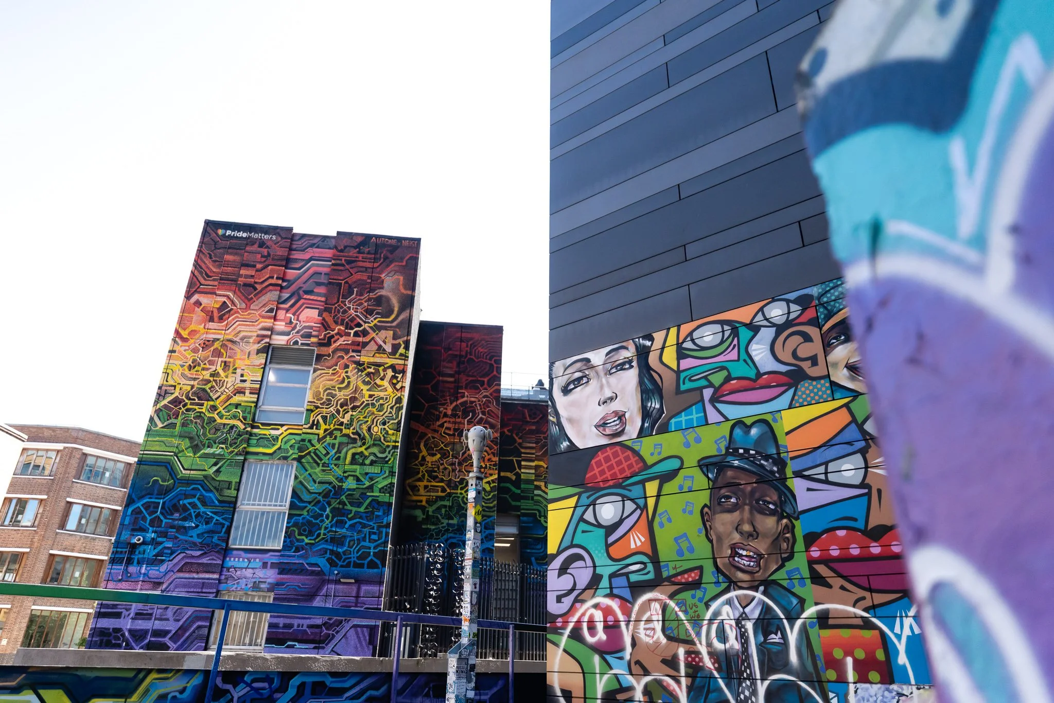 Colorful mural on the side of a building depicting diverse people's faces and abstract designs, with additional murals on neighboring buildings and a clear sky overhead.