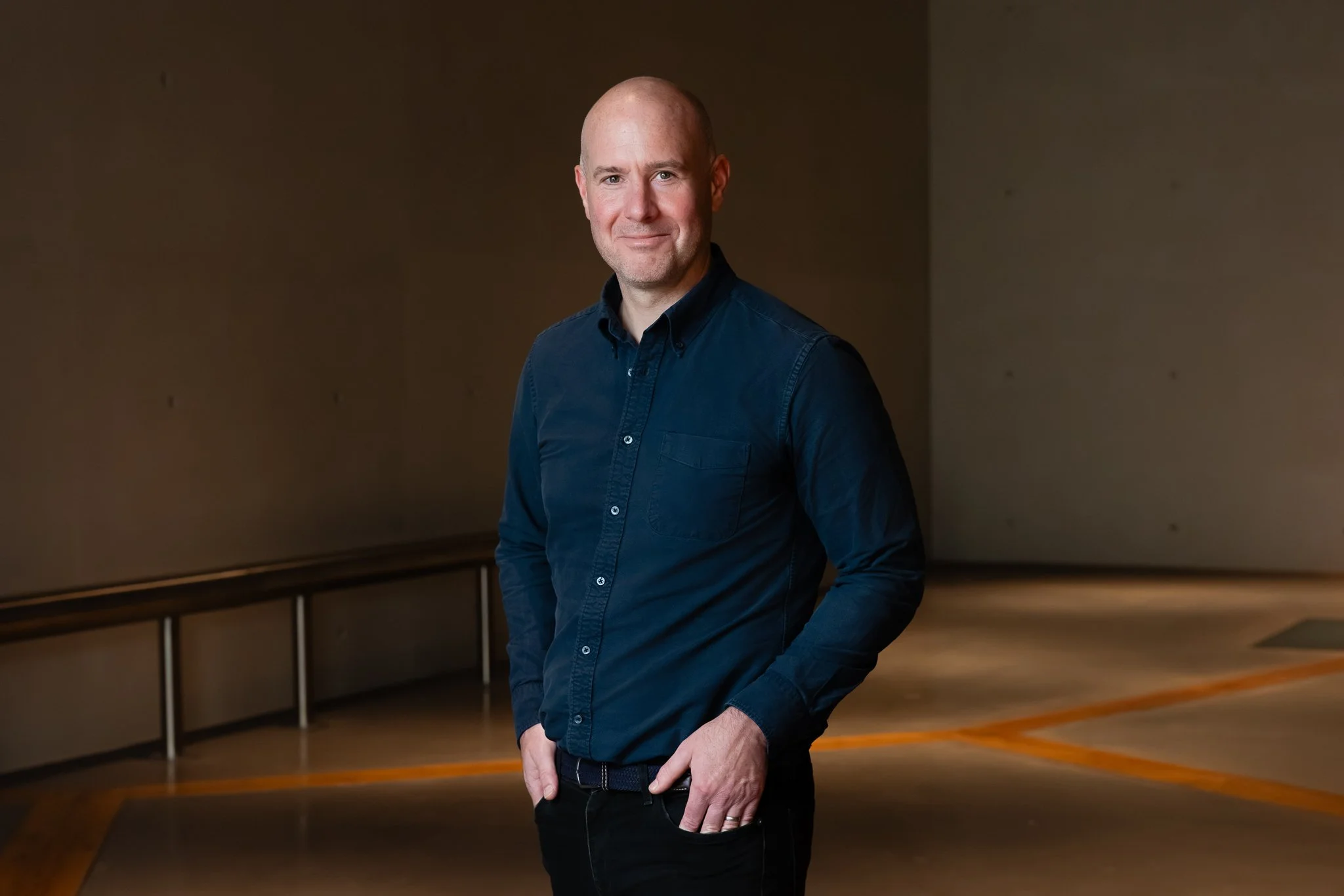 A man with a shaved head and light beard wearing a navy blue button-up shirt standing indoors against a plain background.