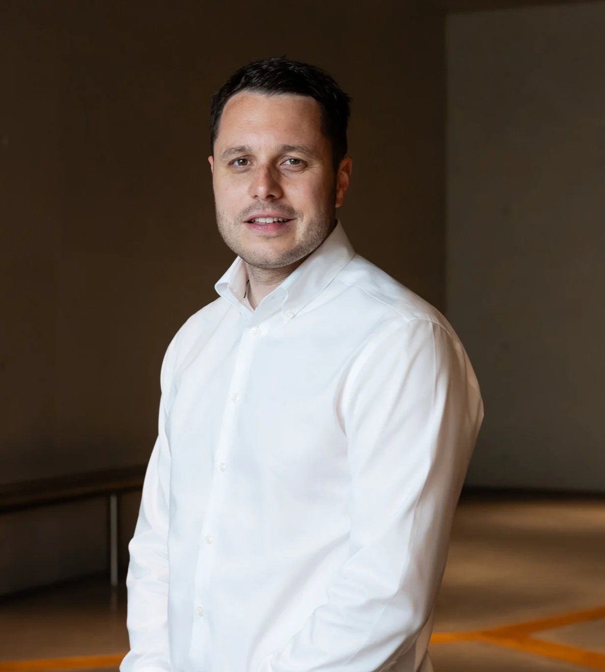 A man with short dark hair, dressed in a white shirt, smiling slightly and looking at the camera. He is standing in an indoor setting with a plain background and wooden furniture behind him.