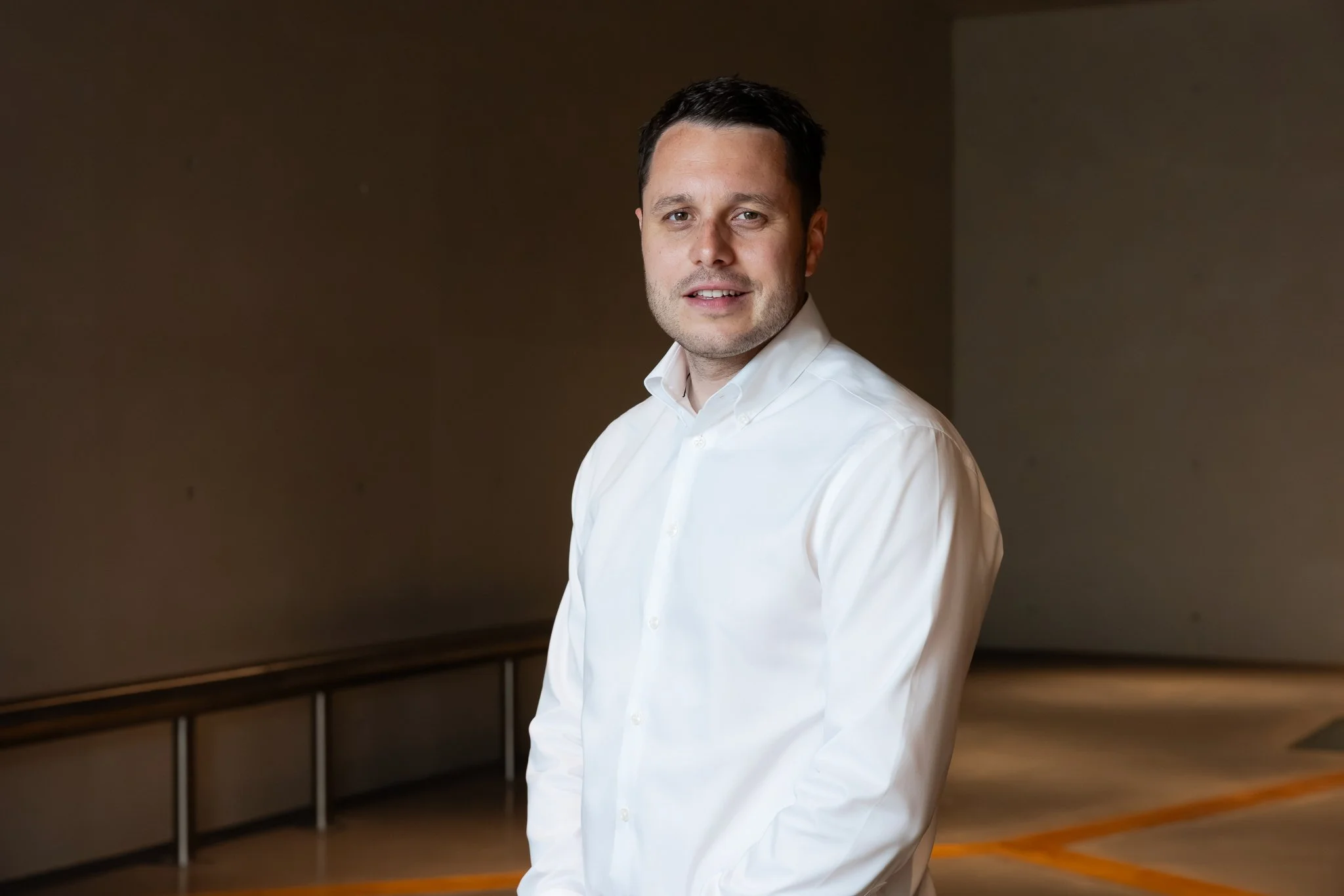 A man in a white shirt standing in an indoor space with a dark background and a railing.