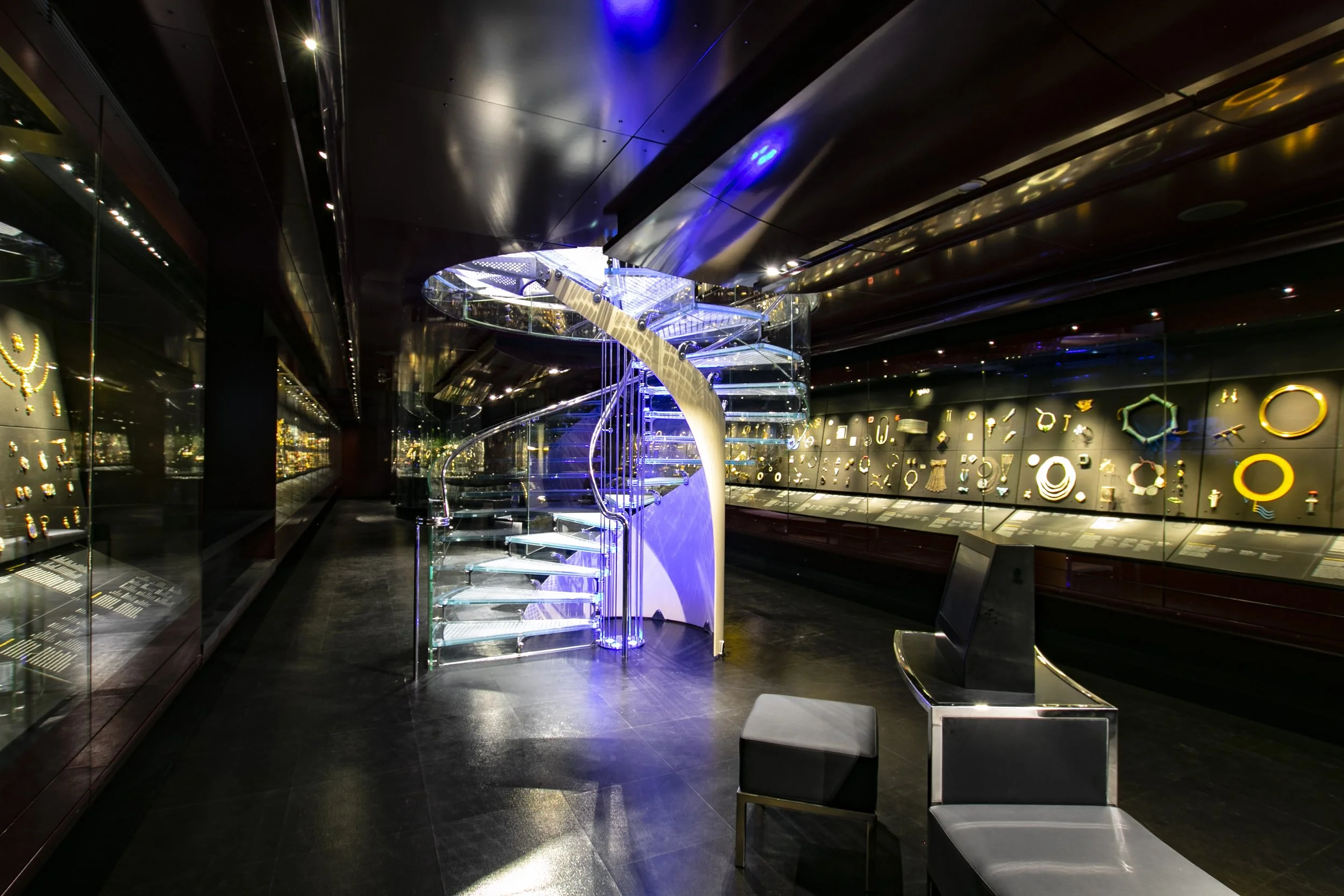 A modern interior space with a glass spiral staircase illuminated with blue lights. The walls are decorated with various illuminated geometric shapes and symbols, and the ceiling has reflective black panels. There are two gray cushioned chairs in the foreground.