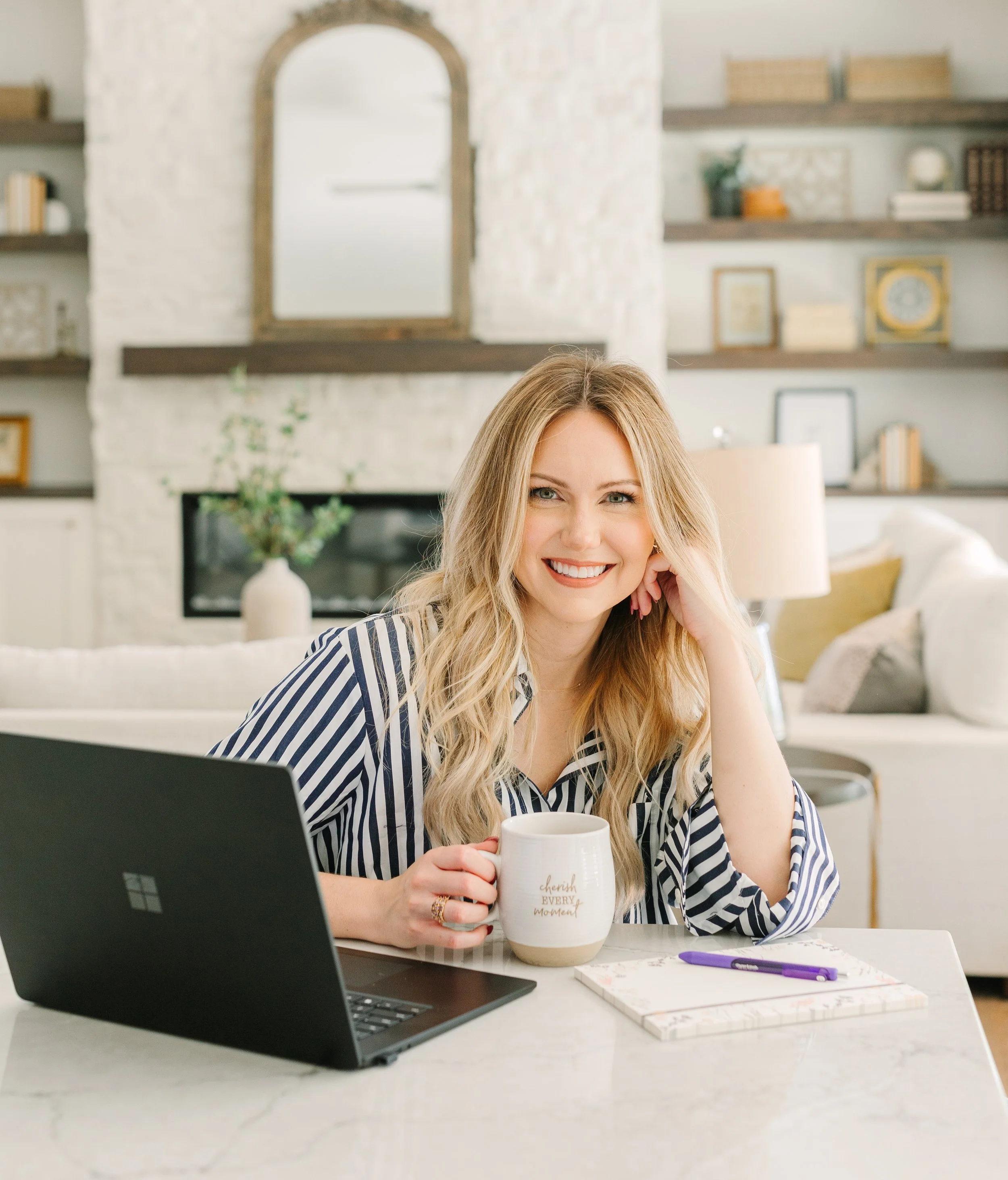 A woman with long blonde hair, wearing a black and white striped shirt, sitting at a white marble desk in a bright, cozy room with shelves and artwork. She is smiling, holding a white mug with gold lettering, and has a notebook and purple pen in front of her. A laptop is partially visible on the left.